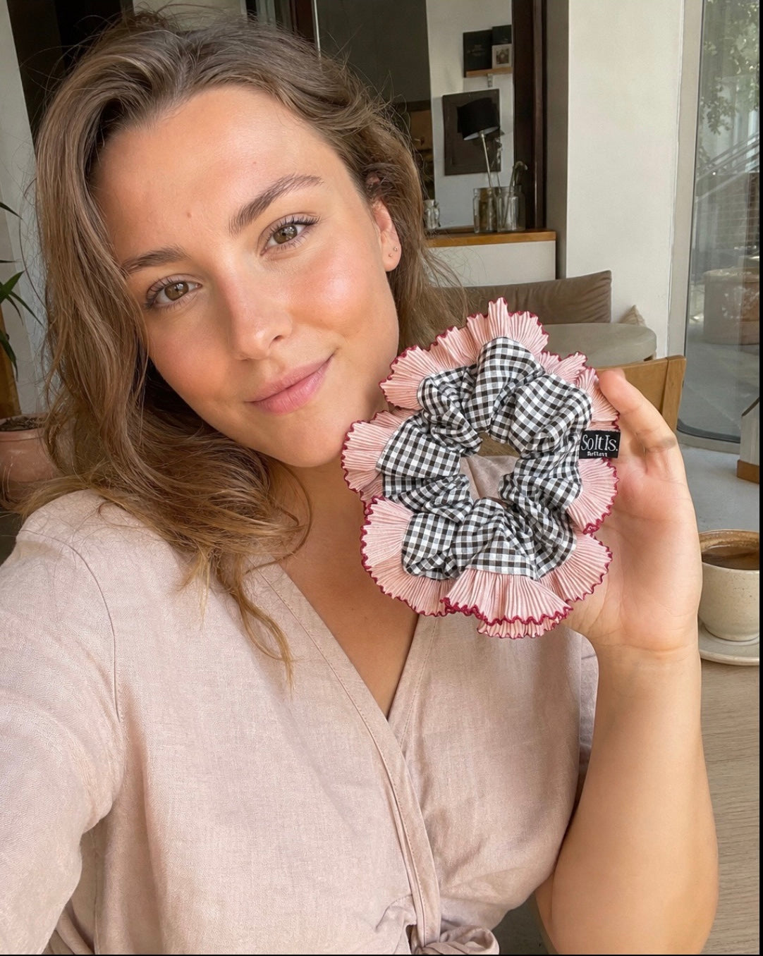 Hand holding an oversized handmade gingham scrunchie with blush pink pleated ruffle edging, styled indoors in natural light.
